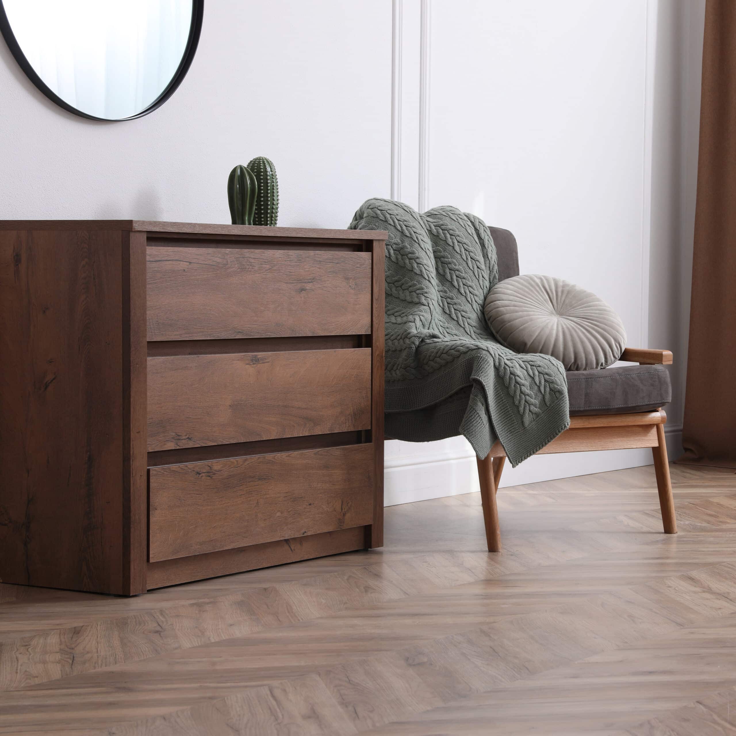 Commode en bois sombre à trois tiroirs, surmontée d'un cactus. Fauteuil gris avec plaid tricoté vert et coussin rond. Parquet chevrons.