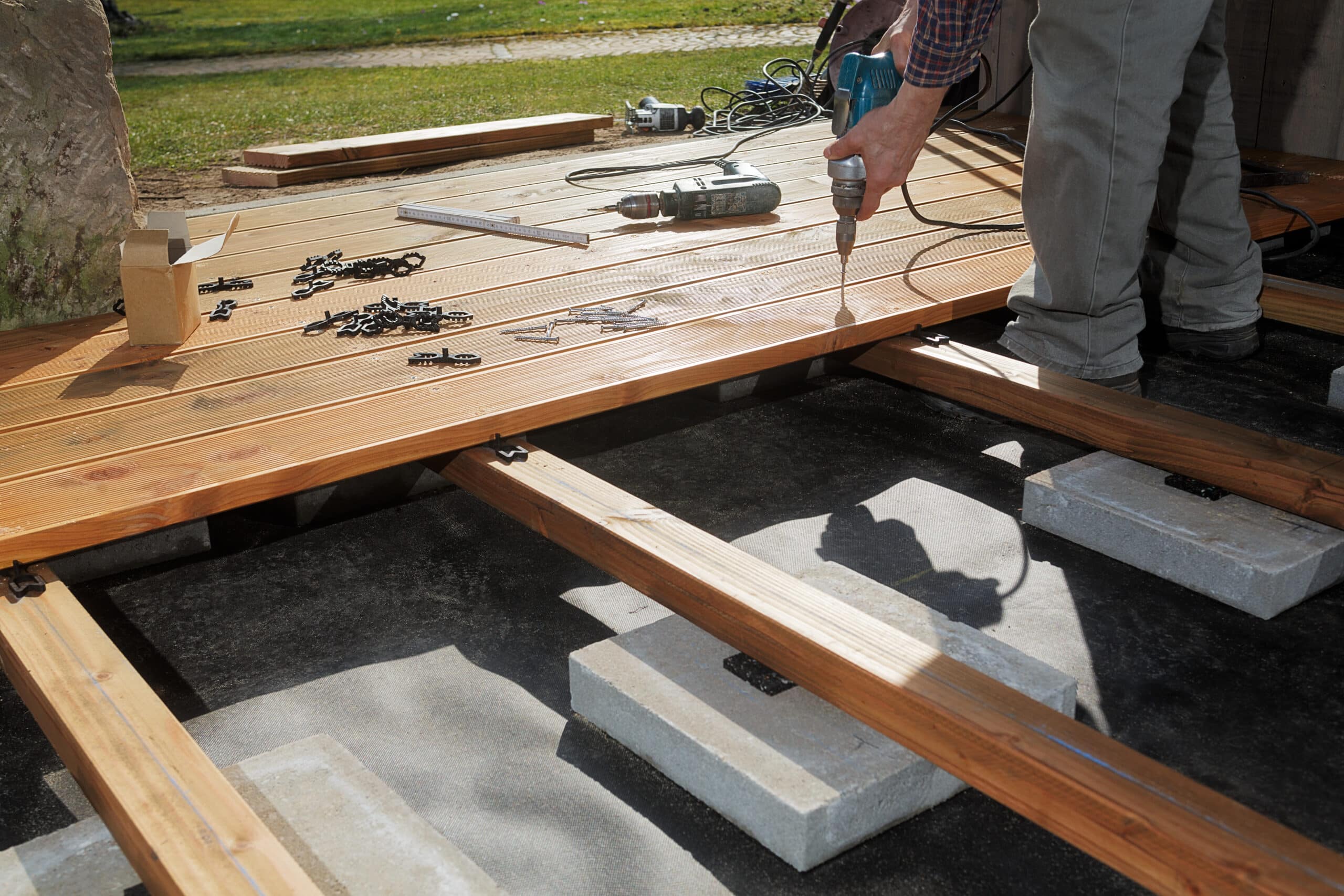Construction d'une terrasse en bois: une personne perce une lame de terrasse sur des lambourdes posées sur des blocs. Outils, vis et clips sont visibles.