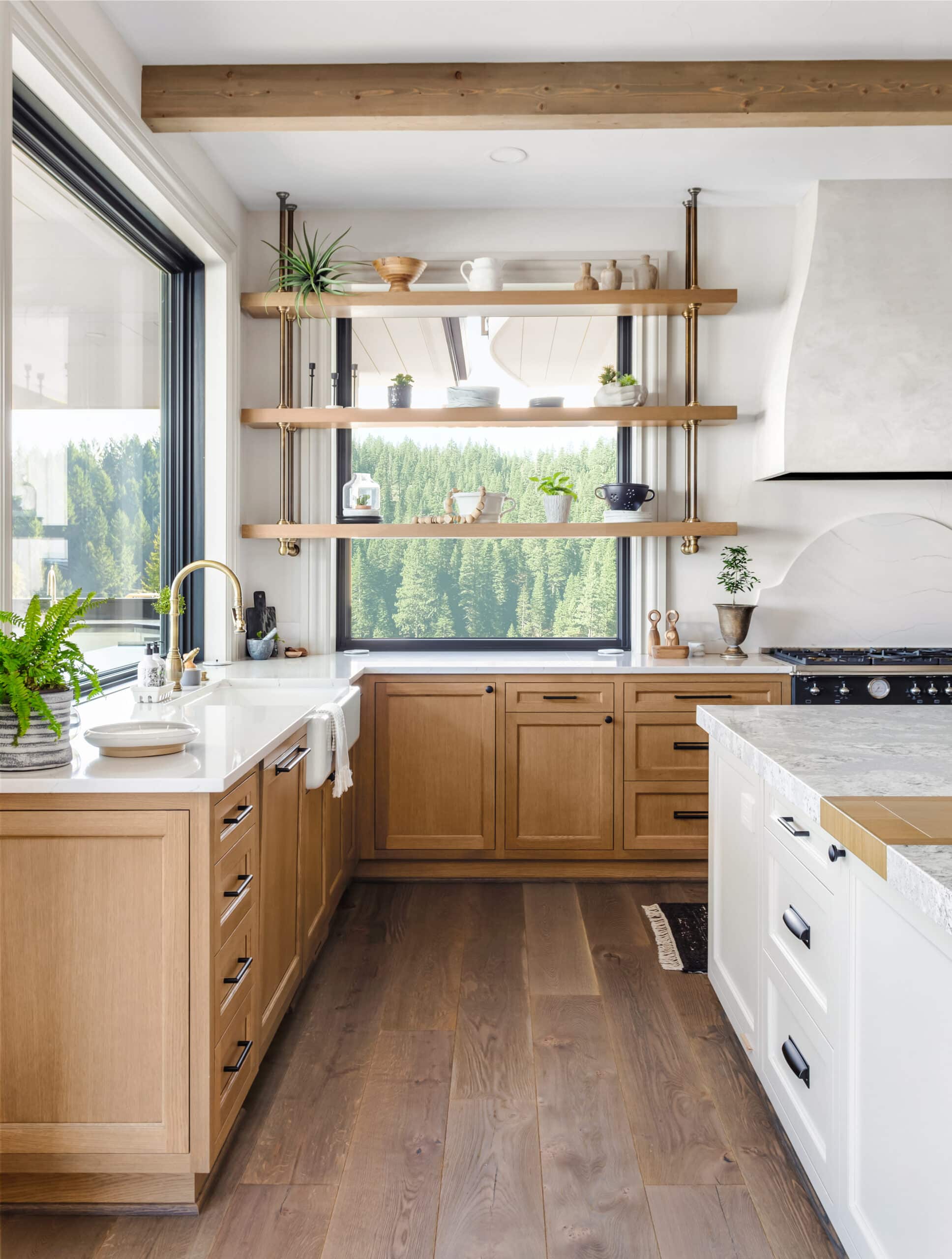 Cuisine moderne lumineuse avec armoires en bois clair, comptoirs blancs, grande fenêtre donnant sur une forêt dense. Étagères ouvertes en laiton.