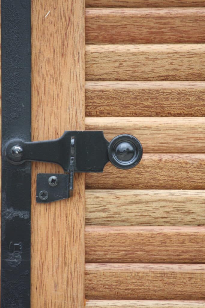 Gros plan sur un loquet noir en métal forgé, fixé sur des lattes de bois clair avec grain apparent. Bande noire texturée avec un logo de serrure stylisé.