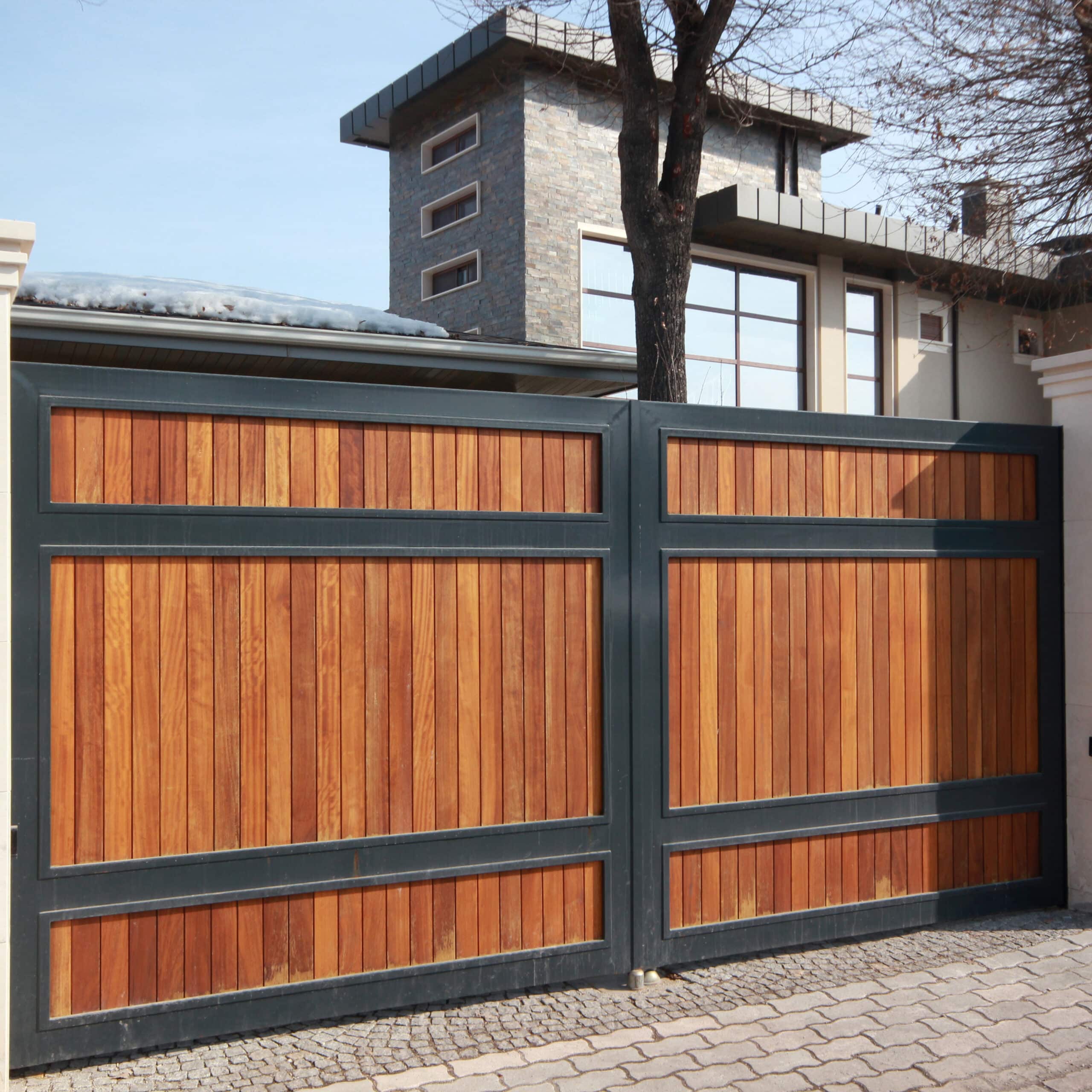 Grand portail moderne en bois clair et métal anthracite devant une maison contemporaine à façade en pierre et grandes baies vitrées. Neige sur le toit.