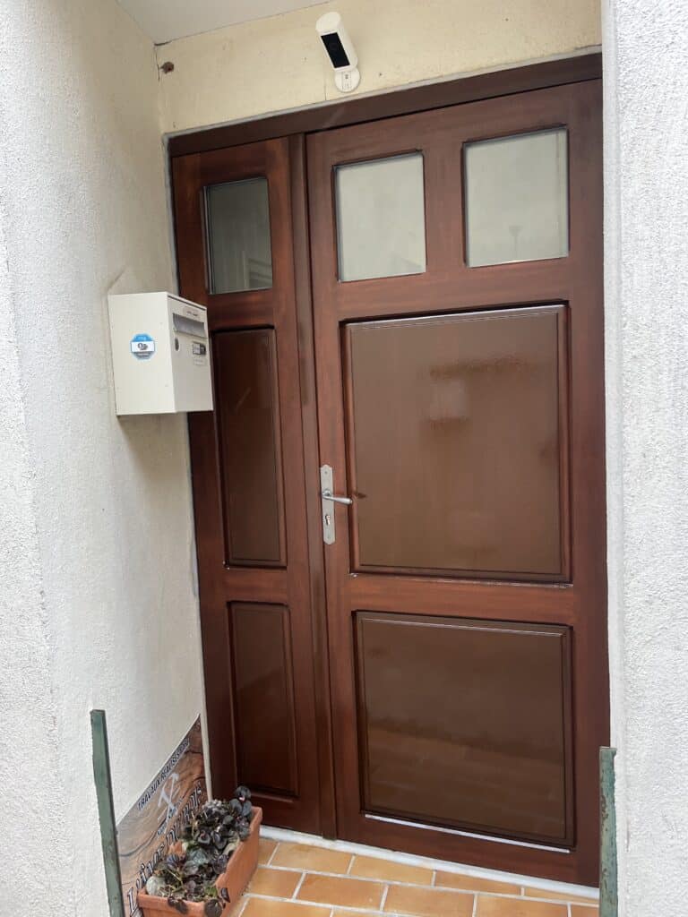 Porte double d'entrée en bois marron brillant avec panneaux vitrés supérieurs. Caméra de sécurité Ring et boîte aux lettres blanche sur le mur.